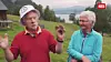 To eldre personer står og prater på en bratt, grønn fjellgard med fjorden i bakgrunnen.