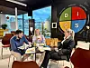 Tre personer sitter ved et rundt bord med papirer og kaffekopper i et moderne møterom.