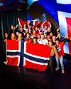 En stor gruppe står samlet innendørs og holder to store norske flagg mens flere løfter hendene.