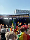 Menneskemengde foran en utendørs scene med skiltet «Ålesund Pride» og regnbueflagg i luften.