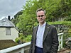 En mann står ved et rekkverk foran et hus og en grønn, bratt skråning.