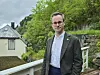 En mann står ved et rekkverk foran et hus og en grønn, bratt skråning.