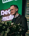 En mann i briller står ved en talerstol med planter foran, foran en grønn bakgrunn med delvis synlig tekst.
