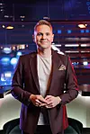 En mann i dress står foran en opplyst bakgrunn i et TV-studio.