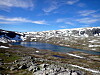 Utsikt over snødekte fjell, en innsjø og steinete høyfjellslandskap under delvis skyet himmel.