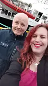 To personer tar selfie foran et cruiseskip ved kai, med en rød livbåt og havneområdet i bakgrunnen.