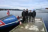 Fire personer står på en brygge ved siden av en blå robåt med norsk flagg.