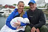 Et par sitter ved en småbåthavn og holder en sovende baby innsvøpt i et teppe.
