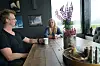 To personer sitter ved et trebord innendørs, med kaffekopper, blomster og utsikt gjennom store vinduer.