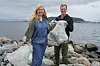 To personer står på en steinete strand med en stor pose med avfall og havet i bakgrunnen.