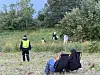 Flere personer i refleksvester står og går i et grønt område med busker og høyt gress.