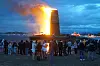 Et stort bål brenner ved sjøen med en folkemengde i forgrunnen og båter i bakgrunnen.