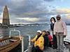 Fire personer står på en brygge ved vannet, med et høyt bålbygg i bakgrunnen.