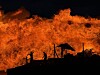 Store oransje flammer stiger opp bak en mørk silhuett av mennesker og strukturer ved vannkanten.