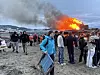 Folk står samlet på en strand mens et stort bål flammer opp i bakgrunnen.