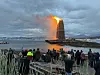 Et stort bål brenner ved fjorden mens mange mennesker ser på fra stranden i skumringen.