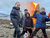 Fire barn står på en steinete strand foran et stort, brennende bål ved sjøen.
