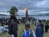 En stor folkemengde står langs strand og fjære og ser et høyt bål brenne under overskyet himmel.