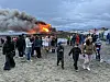 Folk står og går på en strand mens et stort bål brenner kraftig ved fjorden under en overskyet himmel.