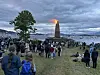 En stor folkemengde står på en gressbakke og langs stranden mens et brennende tårn lyser opp fjorden.