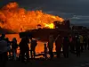 Store flammer fra et bål ved vannet mens publikum står i silhuett langs strandkanten.