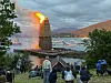 Et høyt bål står i flammer ved sjøen mens folk sitter og står i skråningen og ser på.