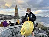 En jente holder en stor gul sekk på en brygge med folk sittende ved vannet og et høyt trebål i bakgrunnen.