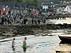 Folkemengde står langs strandkanten ved fjorden, med to personer som bader i forgrunnen.