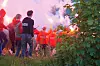 En gruppe mennesker står på gresset og ser på en stor ild og røyk ved vannet.