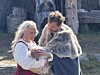 To personer i historiske klær holder et spedbarn foran et tømmerbygg utendørs.