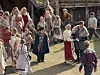 En gruppe mennesker i historiske klær står og går på et utendørs område med trebygninger og steintrapp.