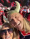 To smilende tilskuere i forgrunnen foran en tettpakket tribune med røde og hvite supportere.