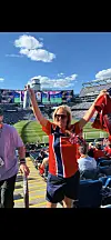En kvinne i rød og svart står på tribunen med armene hevet på et stadion.