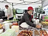 Person som serverer mat ved en utendørs bod med grønnsaker, sauser og grillspyd under et hvitt telt.