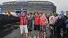 Fire personer i røde og hvite klær står foran et stort stadion i regnvær.
