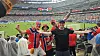 Tre supportere jubler på tribunen foran en fullsatt fotballstadion.