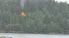 Flammer fra en hytte i skogkledd terreng ved vannet.