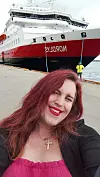 En smilende person med rødt hår står foran et stort cruiseskip ved kaia.