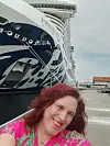 En kvinne smiler i forgrunnen foran baugen på et stort cruiseskip ved kai.