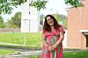 En kvinne i rosa og grønn mønstret kjole står utendørs ved en bygning og smiler mot kamera.