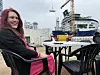 En kvinne sitter ved et utemøbel ved havnen med skip i bakgrunnen.