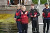 Fire barn står på en brygge ved vannet, iført redningsvester, med fiskestenger og en liten båt i bakgrunnen.