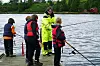Fire barn med fiskestenger står ved vannet sammen med en instruktør i gul jakke.