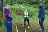 Tre personer står ved et lite kastespill på gresset i en skoglysning.