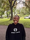 En smilende person i svart hettegenser står i en grønn park med store trær.