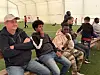 Flere personer sitter på benker inne i et lyst telt med kunstgressbane og følger en kamp på skjerm.