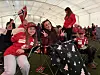 Barn og voksne i røde klær sitter i et telt og følger en fotballkamp mens norske flagg henger i bakgrunnen.