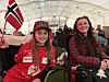 To smilende supportere sitter på stoler i et telt med norske flagg og røde klær.