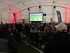 Mange personer sitter i et telt og ser på en fotballkamp på en stor skjerm.