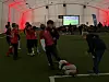 Barn i fotballdrakter står og ser på en kamp på storskjerm i et telt.